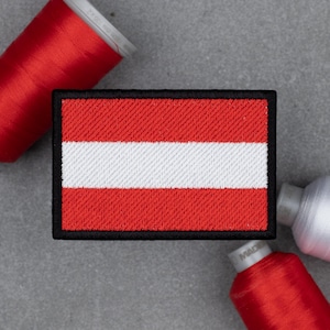 May include: Rectangular embroidered patch displaying the Austrian flag, with red and white stripes and a black border. The patch is set on a grey surface, alongside red and white spools of thread.