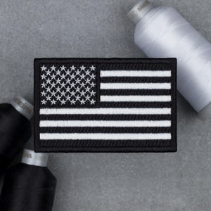 May include: A black and white embroidered patch featuring the American flag design. The patch has a rectangular shape with a black border. The flag design includes white stars in a blue field and white stripes.