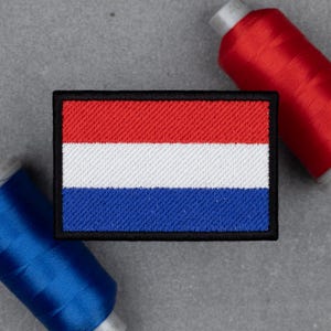 Puede incluir: Parche bordado con la bandera de los Países Bajos. El parche rectangular tiene franjas horizontales rojas, blancas y azules, delineadas con un borde negro. Dos carretes de hilo, uno rojo y otro azul, están en el fondo.