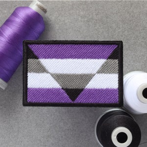 Puede incluir: Un parche bordado con un diseño de triángulo estilizado en morado, blanco, gris y negro. El parche está rodeado por un borde negro.