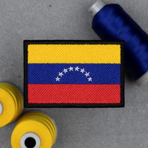 Puede incluir: Parche bordado con la bandera de Venezuela. El parche rectangular tiene una parte superior amarilla, un centro azul con estrellas blancas y una parte inferior roja. El parche está bordeado en negro. Bobinas de hilo amarillo y azul en el fondo.
