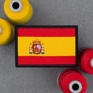 Puede incluir: Un parche rectangular bordado con la bandera de España, que presenta franjas horizontales rojas y amarillas y un escudo central. El parche tiene un borde negro grueso y está rodeado por varios carretes de hilo de coser amarillo y rojo.