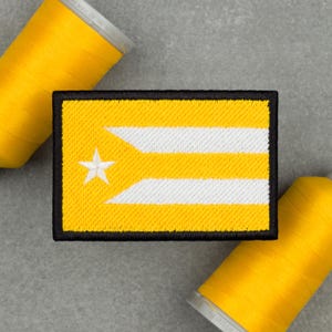 Può includere: Toppa ricamata con un disegno a bandiera gialla e bianca e una stella. La toppa rettangolare ha un bordo nero. Due rocchetti di filo giallo sono sullo sfondo.