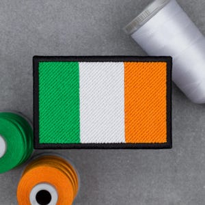 May include: Embroidered patch featuring the flag of Ireland, with vertical stripes of green, white, and orange, framed by a black border. Spools of green, orange, and white thread are visible in the background.