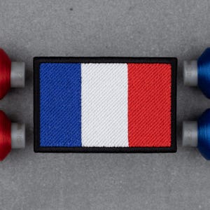 May include: A rectangular patch featuring the French flag, with vertical stripes of blue, white, and red, outlined in black. The patch is surrounded by red and blue spools of thread, suggesting a sewing or crafting theme.