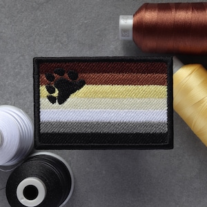 Puede incluir: Un parche bordado marrón, beige, blanco, gris y negro con una huella de pata de oso negra en la esquina superior izquierda. El parche está rodeado por un borde negro.