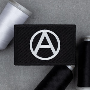 May include: Black rectangular patch featuring a white encircled letter "A" symbol. The patch is made of a textured fabric. White and black spools of thread are in the background.
