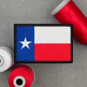 Puede incluir: Un parche rectangular bordado con el diseño de la bandera del estado de Texas. El parche presenta una franja vertical azul a la izquierda con una estrella blanca, y franjas horizontales blanca y roja a la derecha, todo rodeado por un borde negro.