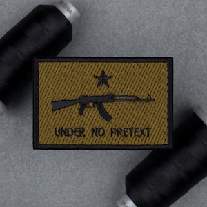 May include: A rectangular embroidered patch with a black border, featuring a dark olive green background with diagonal stitching. It displays a black silhouette of a rifle, a five-pointed star above it, and the text UNDER NO PRETEXT below.