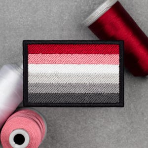 Pode incluir: Patch bordado retangular com um design de bandeira com listras horizontais vermelhas, rosa, brancas, cinzentas e cinzentas escuras, contornado em preto. O patch tem uma aparência texturizada. Bobinas de linha vermelha, branca e rosa no fundo.