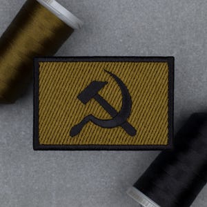 Pode incluir: Patch bordado com um símbolo de martelo e foice preto sobre um fundo dourado e uma borda preta. O patch tem uma textura listrada diagonal. Duas bobinas de linha, uma verde-oliva e outra preta, estão no fundo.