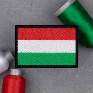 May include: Embroidered patch featuring the Hungarian flag, with horizontal stripes of red, white, and green, framed by a black border. The patch is rectangular and has a textured appearance. Spools of red, white, and green thread are in the background.
