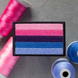May include: A black and white embroidered patch with a six-striped flag design. The flag has pink, purple, blue, and light blue stripes. The patch is surrounded by spools of pink, blue, and light blue thread.