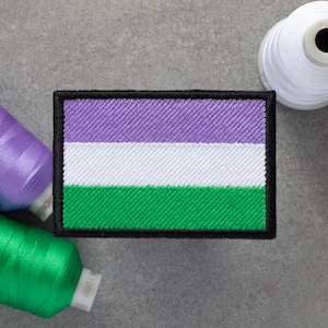 May include: A purple, white, and green embroidered patch with a black border. The patch is a representation of the genderqueer pride flag.