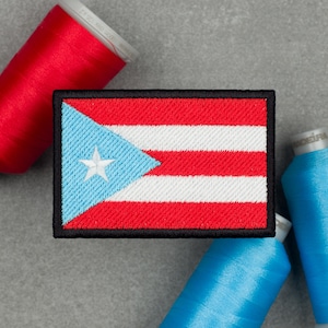 Puede incluir: Parche bordado con la bandera de Puerto Rico. El parche rectangular tiene un borde negro y muestra el diseño de la bandera: una estrella blanca en un triángulo azul, con rayas rojas y blancas. Bobinas de hilo rojas y azules en el fondo.