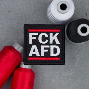 Op de afbeelding: Een vierkante geborduurde patch met een zwarte achtergrond, voorzien van de witte tekst FCK AFD in het midden, omrand door twee horizontale rode strepen aan de boven- en onderkant, omringd door klossen rood, wit en zwart naaigaren.