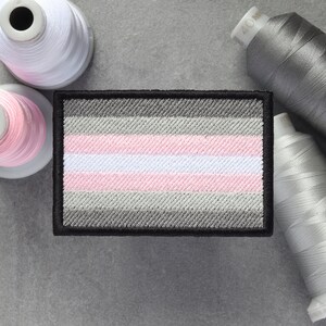 Puede incluir: Un parche bordado con un borde negro, que presenta un patrón de rayas horizontales en tonos de gris, blanco y rosa. El parche se asemeja a una bandera.