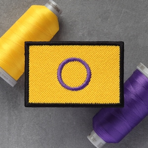 Op de afbeelding: Een gele geborduurde patch met een paarse cirkel erin. De patch heeft een zwarte rand en wordt omringd door gele en paarse spoelen garen.