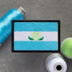 May include: A blue, white, and green embroidered patch with a white rose and green leaves in the center. The patch is surrounded by a black border.