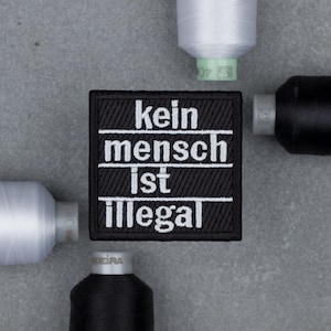 Puede incluir: Un parche cuadrado bordado en negro con el texto blanco "kein mensch ist illegal" dispuesto en cuatro líneas separadas por finas rayas horizontales blancas, rodeado por cuatro carretes de hilo de coser blanco y negro.