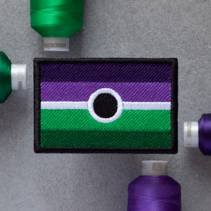 Può includere: Una toppa ricamata in bianco e nero con un design a strisce viola, bianco e verde. La toppa presenta un cerchio nero al centro della striscia bianca.