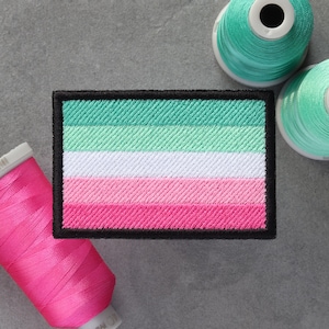 Op de afbeelding: Een groene, witte, roze en lichtroze gestreepte stoffen patch met zwarte stiksels rond de randen. De patch ligt op een grijs oppervlak met spoelen garen.