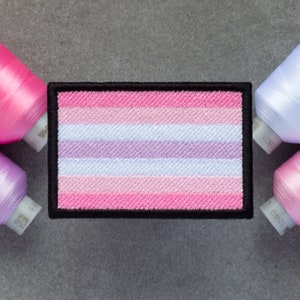 May include: A pink, white, and lavender striped embroidered patch with a black border. The patch is on a gray surface.