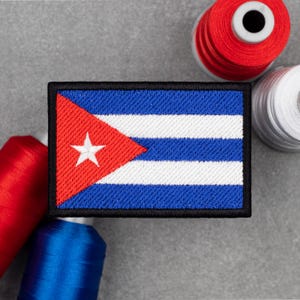 Peut inclure: Un écusson rectangulaire représentant le drapeau cubain, avec un triangle rouge et une étoile blanche à gauche, et des rayures bleues et blanches à droite. Bordure noire. Des bobines de fil rouge, blanc et bleu sont en arrière-plan.