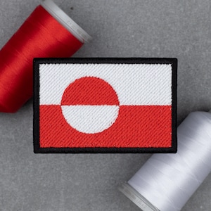 May include: A rectangular patch featuring the flag of Greenland, with a red and white design and a black border. The flag has a white circle on a red background, and a red circle on a white background. Two spools of thread are in the background.