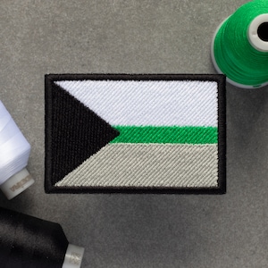 Puede incluir: Un parche bordado en negro, blanco, verde y gris con un triángulo negro en la esquina superior izquierda. El parche tiene un borde negro.