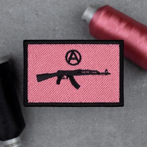 May include: Pink embroidered patch with a black border. The patch features a black AK-47 rifle and an encircled letter "A" on a pink background. The patch is rectangular and appears to be made of fabric.