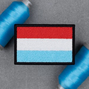 Könnte beinhalten: Gestickter Aufnäher mit der Flagge Luxemburgs, mit horizontalen Streifen in Rot, Weiß und Hellblau, umrandet von Schwarz. Zwei Spulen blauen Garns sind im Hintergrund zu sehen. Der Aufnäher ist rechteckig.