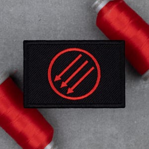 May include: Black rectangular patch with a red circle and three downward-pointing arrows. The patch is made of a textured fabric. Two spools of red thread are in the background.
