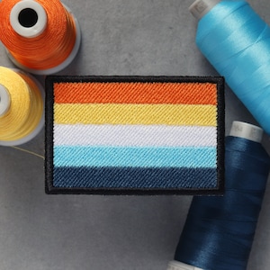 Puede incluir: Un parche bordado colorido con seis rayas horizontales en naranja, amarillo, blanco, azul claro, azul oscuro y negro. El parche está rodeado por un borde negro.