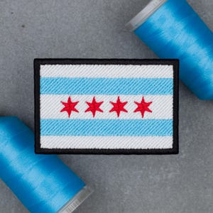 May include: A rectangular embroidered patch featuring the Chicago flag design with white and light blue horizontal stripes and four red six-pointed stars centered in the middle, framed by a thick black border, positioned between two blue thread spools.