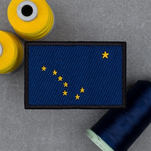 Könnte beinhalten: Ein rechteckiger Aufnäher mit der Flagge von Alaska. Er hat einen dunkelblauen Hintergrund mit acht gelben Sternen. Der Aufnäher ist mit einem schwarzen Rand versehen. Zwei Spulen gelben Fadens und eine Spule blauen Fadens befinden sich im Hintergrund.