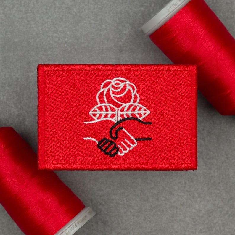 An embroidered patch with a red background featuring a white rose and two hands shaking, one outlined in white and the other in black. The design symbolizes democratic socialism. The patch has a red border and is displayed on a gray surface, surrounded by red spools of thread