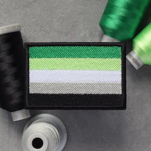May include: An embroidered patch with a flag design in green, light green, white, grey, and black. The patch is surrounded by spools of thread in black, green, and grey.