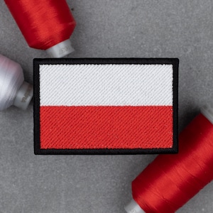 Puede incluir: Un parche rectangular con la bandera polaca, con una franja blanca sobre una franja roja, contorneado en negro. Carretes de hilo rojo y blanco rodean el parche, lo que sugiere un tema artesanal o de costura.