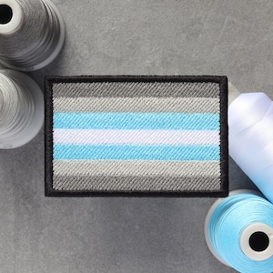 Pode incluir: Um remendo bordado representando a bandeira do orgulho genderqueer. O remendo é cinza, branco e azul claro com uma borda preta.