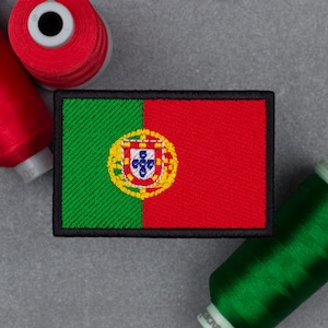 May include: A rectangular embroidered patch featuring the flag of Portugal with a green left section and a red right section, centered with a yellow, red, and white coat of arms. The patch has a thick black border and is surrounded by thread spools.