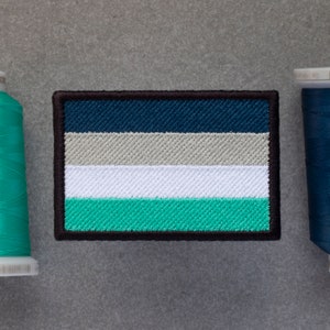 Puede incluir: Un parche de tela a rayas azul, gris, blanco y verde con costuras negras alrededor de los bordes. El parche es una representación de la bandera del orgullo genderqueer.