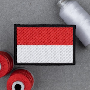 Op de afbeelding: Geborduurde rechthoekige patch met de Indonesische vlag, met een rood bovenste deel en een wit onderste deel, omlijnd door een zwarte rand. Spoelen met rood en wit garen zijn op de achtergrond te zien.