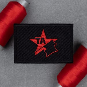 May include: Black rectangular patch with a red star design and the letters "TA" embroidered in the center. The patch is surrounded by two spools of red thread.