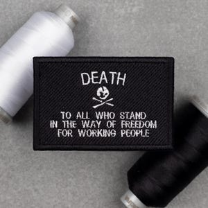May include: Black rectangular patch with white embroidered text. The text reads "DEATH" above a skull and crossbones, and "TO ALL WHO STAND IN THE WAY OF FREEDOM FOR WORKING PEOPLE." White and black thread spools are in the background.