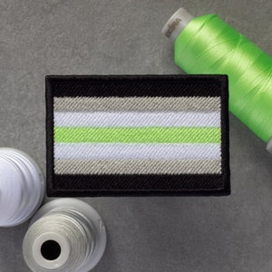 May include: A black, white and light green embroidered patch with a design resembling a pride flag. The patch is surrounded by spools of white and green thread.