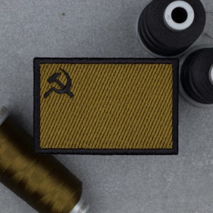 Puede incluir: Un parche rectangular con borde negro e interior dorado con un símbolo negro de la hoz y el martillo. El parche tiene un patrón de rayas diagonales. Bobinas de hilo en el fondo.