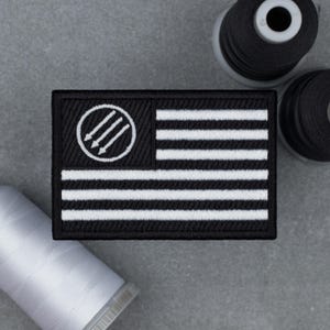 Puede incluir: Parche bordado en blanco y negro de forma rectangular. Presenta un diseño de bandera con rayas blancas horizontales y un campo negro. Un círculo blanco con tres flechas apuntando hacia abajo es visible. Borde negro.