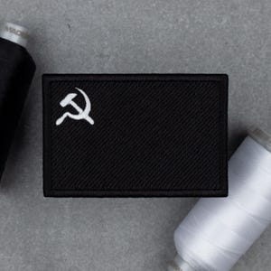 May include: Black rectangular patch with a white hammer and sickle emblem. The patch is surrounded by black thread and sits on a gray surface. Two spools of thread, one black and one white, are also visible.