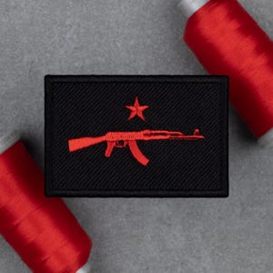 May include: Black rectangular patch with a red AK-47 rifle and a star embroidered on it. The patch is surrounded by red thread spools. The patch is likely for a jacket, backpack, or other apparel.